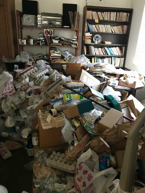 Image shows a living room completely block by trash and clutter. There are empty egg cartons, empty water bottles, food, various bottles, boxes and bags.